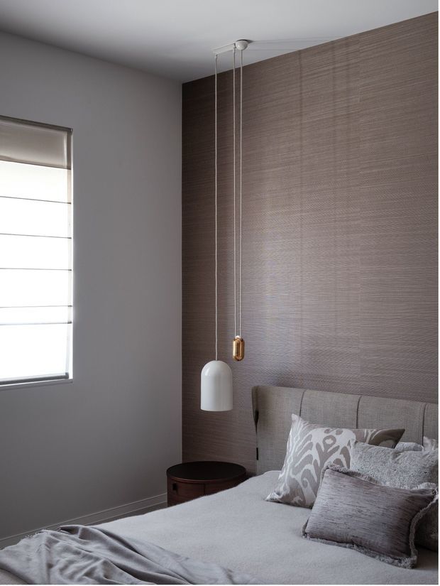 A refined contemporary bedroom with a muted grey and taupe palette. The room features a textured taupe feature wall behind a large upholstered bedhead, layered grey bedding and patterned cushions, a small dark timber bedside table, a white pendant light with exposed cords, and a roller blind over the side window.