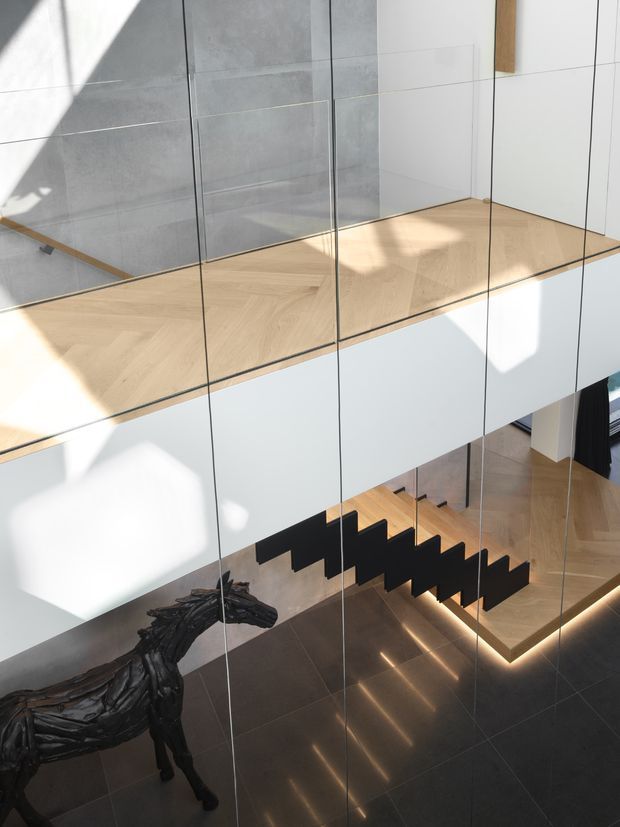 A contemporary multi-level interior viewed through extensive floor-to-ceiling glazing or mirrored panels. The space features a floating black stair with timber treads, pale timber flooring at the upper level, white minimalist wall panels, large grey wall tiles, dark polished flooring below, and integrated linear stair lighting. A sculptural dark horse-like artwork is visible in the lower foyer.