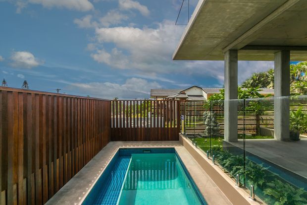 A compact rectangular swimming pool sits within a private modern courtyard, bordered by pale paving and tall vertical timber fencing. To the right is a covered concrete terrace with substantial square columns and frameless glass balustrading, with tropical planting beyond. The image has a bright coastal outdoor atmosphere and shows a neighbouring pitched-roof dwelling in the background.