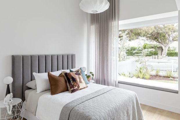 Bright, refined bedroom with a tall vertically channelled upholstered headboard, white bedding, layered neutral and patterned cushions, full-height sheer curtains, pale timber flooring, and a large picture window overlooking a landscaped residential setting. A sculptural pendant light and small bedside furnishings complete the calm contemporary scheme.