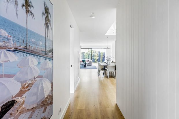 A bright contemporary residential hallway with pale timber flooring and white vertical wall panelling. The corridor opens toward a dining area and living space with large glazed doors or windows at the rear. A large coastal beach-themed artwork occupies the left wall, while recessed ceiling lights and clean-lined finishes create an airy, minimal interior.
