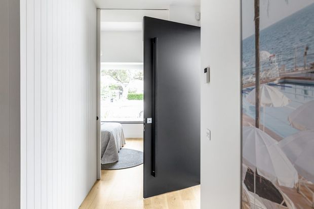 Bright minimalist hallway looking through an open matte-black door into a bedroom. The corridor has white walls, vertical white wall panelling, pale timber flooring, and a large framed artwork on the right. The bedroom beyond contains a partially visible bed, rug, and abundant daylight.