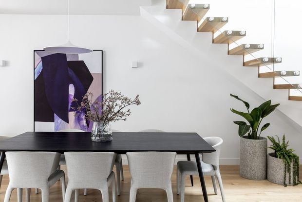 Bright contemporary dining area with a long dark dining table and upholstered dining chairs, set against white walls and pale timber flooring. A large abstract artwork and vase with branches sit behind the table, while an open-riser timber staircase with glass balustrade rises along the right side. Indoor plants add a natural accent.
