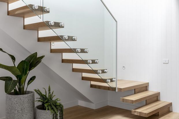Modern minimalist floating timber staircase with open risers, cylindrical metal supports, and a clear glass balustrade. The stair rises beside a white vertically panelled wall, with timber flooring and potted plants beneath.