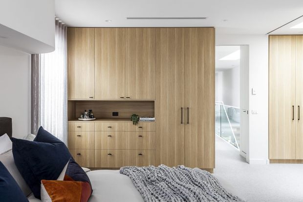 Bright contemporary bedroom with extensive light timber built-in cabinetry, including a wardrobe and integrated storage unit with a recessed display niche. A bed with layered navy and rust cushions and a chunky grey throw occupies the foreground. The room opens toward a bright stair or landing area, with soft carpet flooring and sheer curtains.