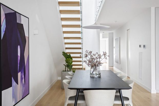Bright contemporary open-plan dining area with a large dark dining table and upholstered chairs, positioned beneath a floating timber-and-white stairway. White walls, light timber flooring, minimalist pendant lighting, abstract artwork, indoor plants, and a glazed stair balustrade create a clean, refined interior.