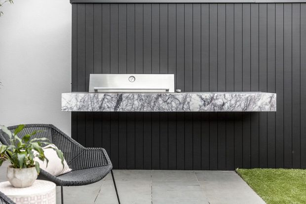 Contemporary outdoor entertaining area featuring a built-in stainless-steel barbecue set into a dramatic veined stone benchtop against a dark vertical-panel feature wall. A woven lounge chair, side table with potted plant, paved terrace and a small lawn area complete the scene.