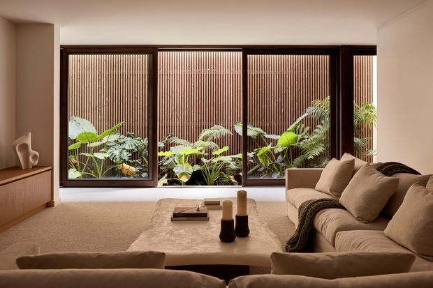 Warm, refined living room with a large neutral sectional sofa arranged around a low stone or concrete coffee table. Floor-to-ceiling dark-framed glazing spans the rear wall, opening visually onto lush planting and a vertical timber screening wall. Built-in timber cabinetry and understated decor complete the calm, contemporary interior.