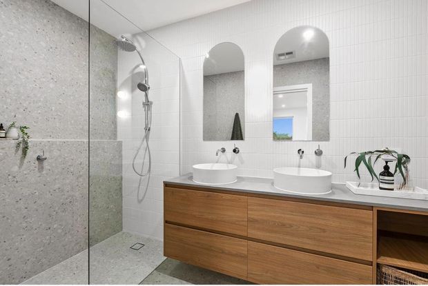 Bright contemporary bathroom with a walk-in shower enclosed by clear glass, pale terrazzo-look wall and floor surfaces, white vertical wall tiles, and a long timber vanity with two white vessel basins. Arched mirrors, wall-mounted taps, recessed lighting, and open shelving complete the clean, refined fit-out.