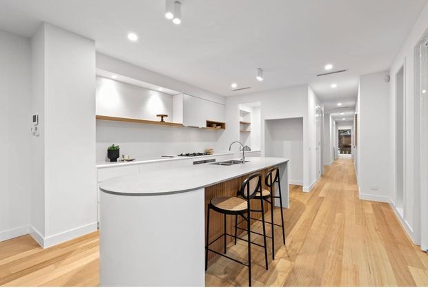 Bright contemporary open-plan kitchen with a curved white island and integrated sink, timber-look cabinetry, open shelving, and three black metal bar stools. The kitchen opens onto a long hallway with light timber flooring and multiple white interior doors. Recessed and track lighting create an airy, clean interior.