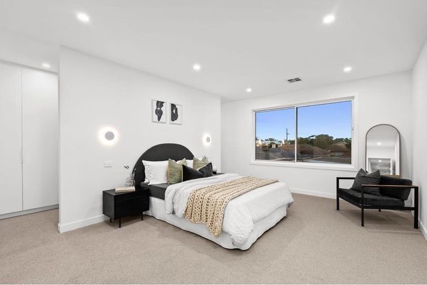 Bright contemporary bedroom with white walls and ceiling, beige carpet, a large upholstered bed with a dark rounded headboard, black bedside table, neutral bedding, and a black lounge chair beside an arched full-length mirror. A wide window provides suburban views and abundant natural light.