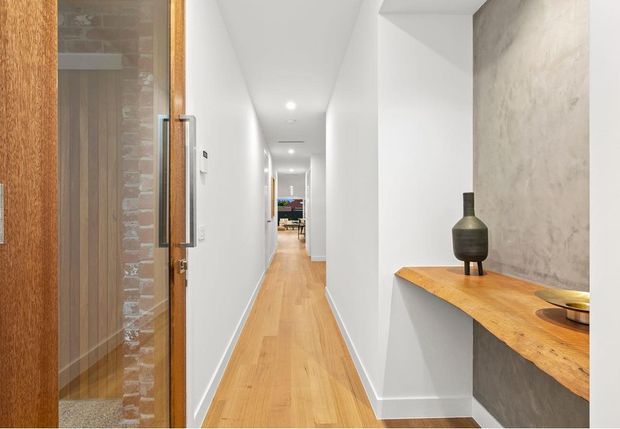 A bright, narrow hallway with light timber flooring and white walls, viewed from a timber-framed glazed entry door. A built-in timber console with a dark textured wall backdrop occupies the right foreground, while a long corridor leads toward a distant living area. Recessed ceiling lighting and clean-lined finishes create a contemporary, uncluttered interior.