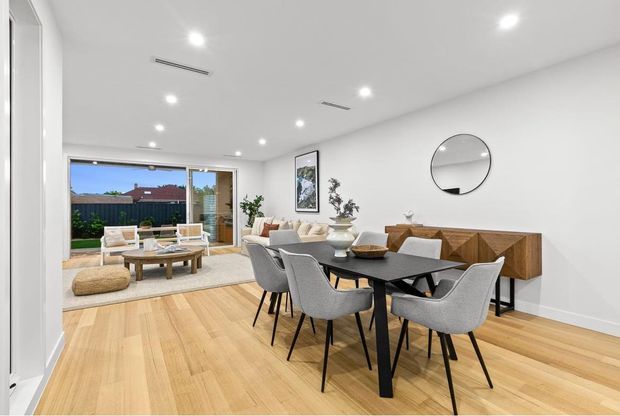 Bright open-plan dining and living area with pale timber flooring, a black dining table surrounded by upholstered chairs, and a sculptural timber sideboard against a white wall. The adjacent living area opens through large sliding glass doors to an outdoor space, with recessed ceiling lighting and minimalist contemporary furnishings.