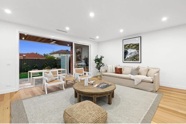 Bright, open-plan living room with white walls and recessed ceiling lights, warm timber flooring, a large neutral rug, cream sofa, woven ottoman, and round timber coffee table. Wide sliding glass doors open to a small outdoor patio and garden, with a framed nature print providing a subtle accent.