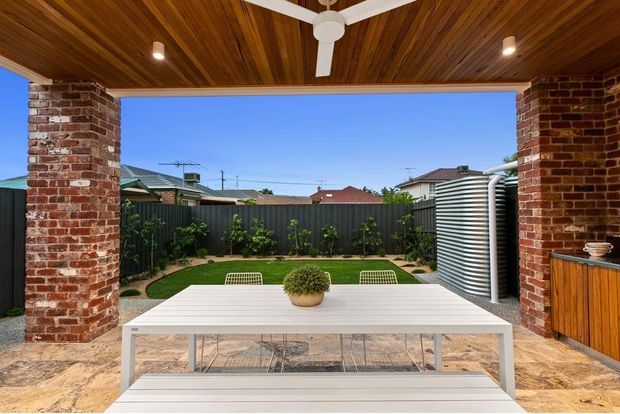 Covered outdoor entertaining area opening onto a landscaped backyard. The space features exposed red-brick columns, a timber-lined ceiling with ceiling fan and downlights, tiled flooring, a large white outdoor dining table, built-in timber cabinetry, and a corrugated metal rainwater tank beside the garden.
