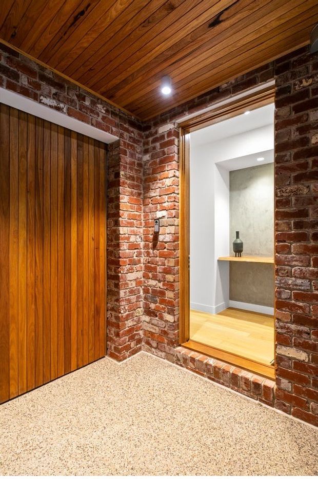 Interior view of a compact entry or hallway with exposed red brick walls, a warm timber-lined ceiling, tall timber-clad built-in doors, and a terrazzo-style speckled floor. A raised opening leads into a brighter adjoining area with timber flooring, a minimalist built-in shelf, and a concrete-look feature wall.