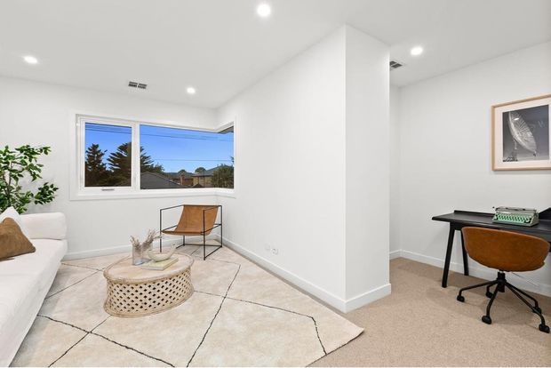 Bright, minimalist open-plan living and home office area with white walls, recessed ceiling lights, large windows, a white sofa, woven round coffee table, neutral rug, and a compact black desk with leather office chair. A partial dividing wall separates the workspace from the sitting area.