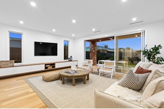 Bright contemporary living room with white walls and ceiling, recessed downlights, light timber flooring, a long low built-in media cabinet, wall-mounted television, woven area rug, round timber coffee table, and a large cream sectional sofa with striped cushions. Wide sliding glass doors open to an outdoor entertaining area and garden, with a brick feature wall visible outside.
