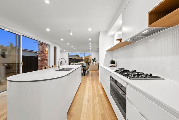 Bright contemporary open-plan kitchen with a long curved white island, integrated sink, white handleless cabinetry, white vertical tile splashback, timber open shelving, stainless-steel appliances, and warm timber flooring. The kitchen opens toward a dining and living area, with large glazed doors and windows providing strong natural light.