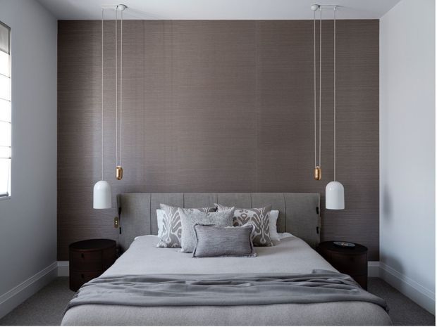 A calm, symmetrical contemporary bedroom with a grey upholstered bed centred against a full-height textured taupe feature wall. Matching dark round bedside tables flank the bed, while paired white pendant lights hang from the ceiling. The room has pale grey walls, grey carpet and layered neutral bedding with patterned cushions.