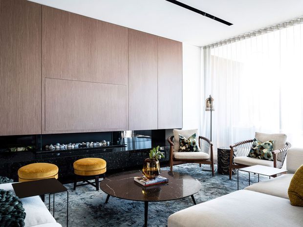 Refined contemporary living room with a large wall of warm wood veneer cabinetry, dark reflective stone or glass lower joinery, integrated fireplace, sculptural coffee table, pale upholstered seating, woven armchairs, and full-height sheer curtains. The room is bright and carefully styled with a muted neutral palette and darker accents.