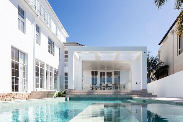 A bright contemporary courtyard-style residence viewed from the swimming pool. The image features a large white multi-storey building with extensive gridded glazing, a covered outdoor entertaining pavilion with a rectangular opening and skylight-like roof apertures, broad steps, pale stone retaining details, and a turquoise pool in the foreground. Palm trees and neighbouring structures are visible around the edges.