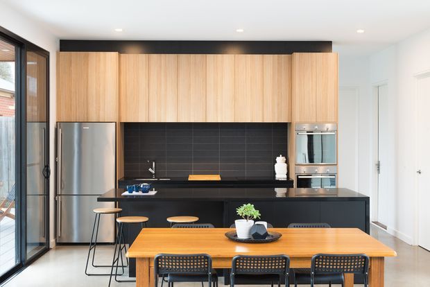 Bright contemporary open-plan kitchen and dining area with full-height timber cabinetry, a black tiled splashback, dark island cabinetry and benchtop, stainless-steel appliances, and a timber dining table with black chairs. Large glazed doors bring in natural light, while white walls and recessed ceiling lights create a clean, modern feel.