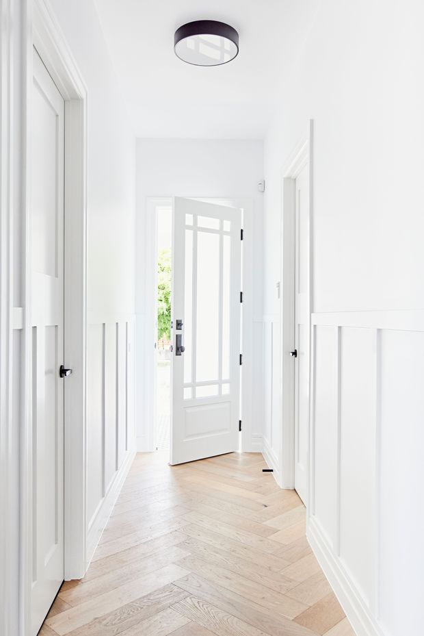 Bright, narrow residential hallway with white paneled walls, multiple white interior doors, decorative wall moulding, a glazed front door opening to greenery, and a pale timber herringbone floor. A dark circular flush-mount ceiling light provides contrast.