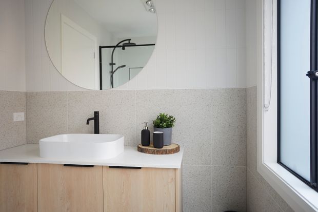 Bright contemporary bathroom featuring a floating timber vanity with a white vessel basin, matte black tapware and hardware, a terrazzo-look tiled splashback, a large round mirror, and a black-framed window.