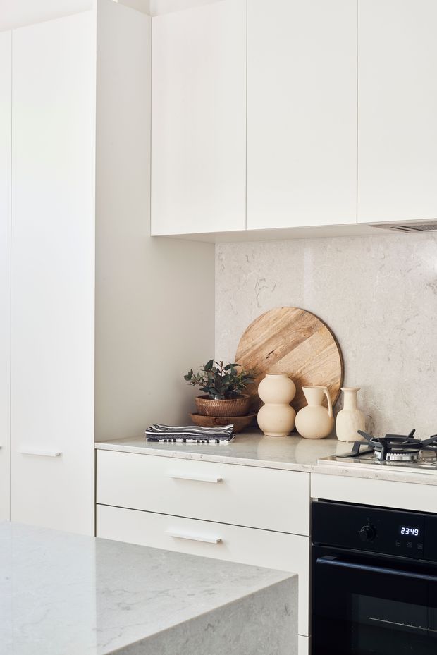 A bright, minimalist kitchen with full-height white cabinetry, a pale stone benchtop and matching stone splashback, integrated black oven and gas cooktop, and a foreground island. Warm timber accessories and neutral ceramic vessels add a subtle natural accent.