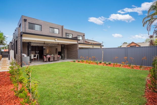 Exterior view of a contemporary two-storey brick house with a large covered outdoor entertaining area opening onto a broad lawn. The home has dark brickwork, horizontal balcony and roof forms, extensive glazing, and a fenced landscaped garden with red mulch and planted borders. A clear blue sky and palm trees are visible in the background.
