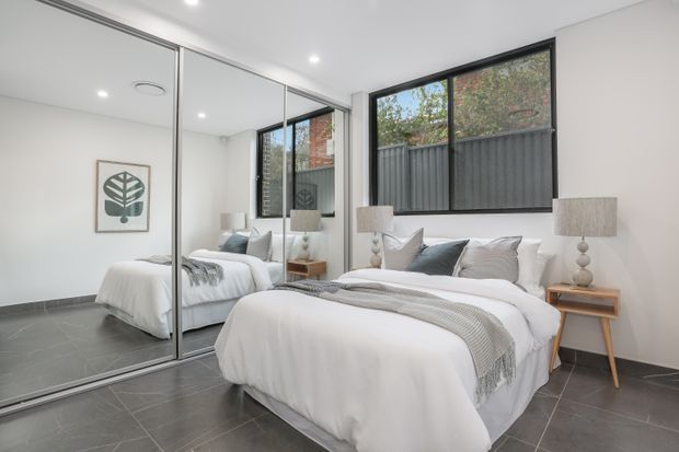 Bright contemporary bedroom with a neatly made bed, white bedding, grey throws and cushions, mirrored built-in wardrobe doors, dark large-format tiled flooring, bedside tables, and matching table lamps. A large black-framed window provides views to a privacy fence and greenery outside. The room has clean white walls, recessed ceiling lights, and a calm neutral palette.