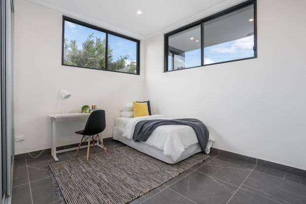 Bright, minimalist bedroom with a single bed, white bedding, grey throw, yellow accent cushion, compact white desk, black chair, and a neutral woven rug. Large black-framed corner windows provide natural light and views of greenery and sky. The room has white walls and ceiling, dark grey tiled flooring, and simple recessed lighting.