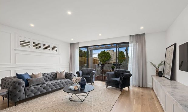 Bright, refined living room with a deep tufted grey sofa, matching armchairs, patterned rug, round coffee table, white decorative wall panelling, timber flooring and a long white media console. Full-height glazed doors open to an outdoor entertaining area, with sheer curtains and recessed ceiling lights creating an airy, polished interior.
