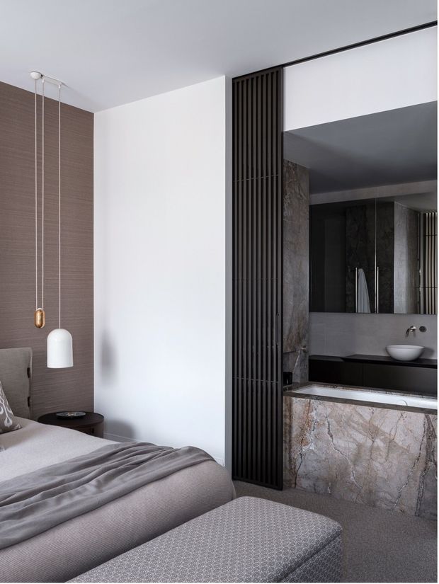 A refined contemporary bedroom with a partially open ensuite bathroom. The bedroom features a soft grey upholstered bed, layered grey bedding, a patterned upholstered bench, a textured taupe wall, and minimalist pendant lighting. A dark vertical slatted screen separates the bedroom from the bathroom, which includes a floating vanity, vessel basin, large mirror, dark cabinetry, and a veined stone-clad bath or vanity surround. The palette is predominantly grey, taupe, white, black, and natural stone, creating a calm and luxurious atmosphere.