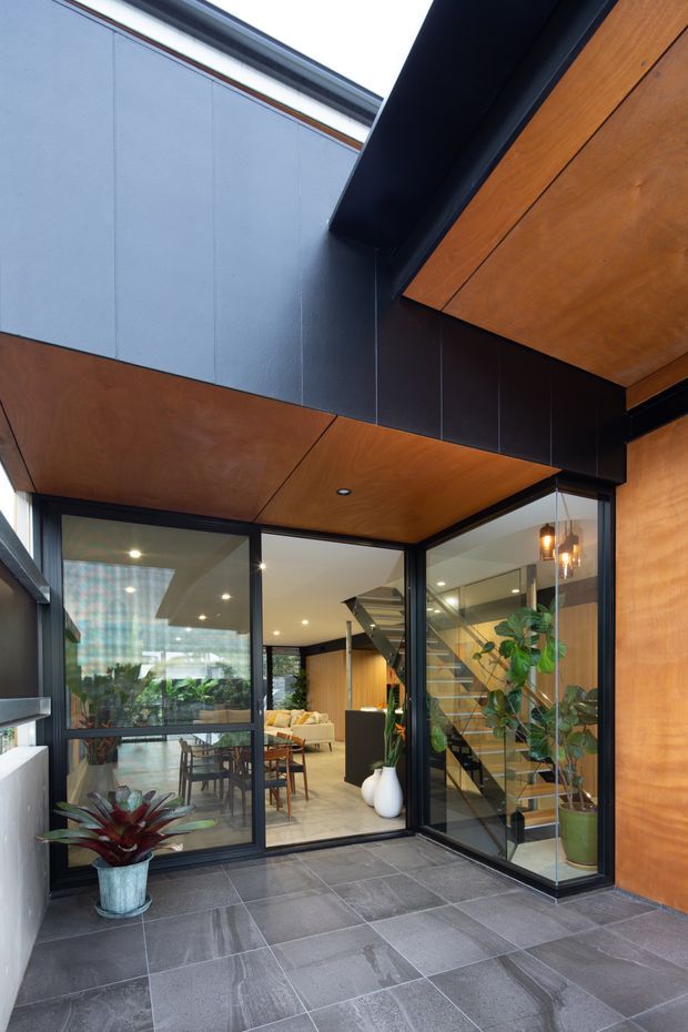A contemporary covered entry courtyard or patio viewed from outside, with large-format grey tiled paving, dark metal-framed floor-to-ceiling glazing, warm timber-lined soffits and wall cladding, and a glazed stairwell visible inside. The glazing opens onto a bright living and dining area with indoor plants, while a black structural beam and crisp geometric lines create a strong modern architectural composition.