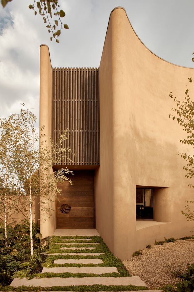 A sculptural residential facade with smooth warm earthen-toned plaster walls, curved parapet forms, and a tall vertical timber screen above a recessed timber entry door. A stepping-stone path crosses low greenery toward the entrance, while a small deep-set window punctuates the side wall. The exterior is photographed in a landscaped setting with soft overcast light and autumn foliage.