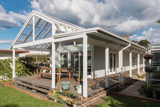 Exterior view of a bright single-storey home extension with a glazed gable-roofed sunroom and covered timber deck. White weatherboard walls, white-painted structural posts, extensive framed glazing, and a low-pitched roof create a light, garden-facing outdoor living area. The deck includes a timber dining table and bench, while brick edging, potted plants, and garden beds surround the structure. Dramatic cloudy sky and a suburban garden form the background.