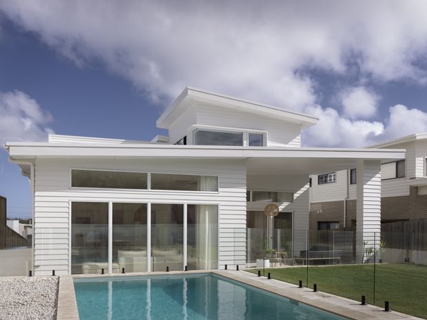 Contemporary white two-storey residence viewed from the rear, featuring horizontal weatherboard cladding, broad glazed openings, a covered outdoor entertaining area, and a rectangular swimming pool in the foreground. The architecture has strong horizontal lines and a low-pitched projecting roof, with neighbouring homes visible beyond the property.