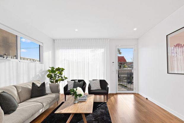 Bright contemporary living room with white walls and ceiling, sheer floor-to-ceiling curtains, timber flooring, a neutral sectional sofa, two dark armchairs, a low timber coffee table, and a black rug. Large windows and a glazed exterior door provide abundant natural light.