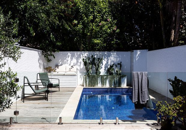 A compact outdoor pool courtyard surrounded by white rendered privacy walls and lush planting. The blue tiled rectangular pool sits beside a timber deck furnished with a lounge chair and built-in outdoor seating, while raised planters and towels contribute to the relaxed residential setting.
