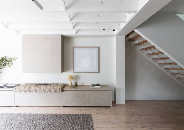 Bright minimalist interior featuring a low built-in storage console, pale textured cabinetry, white walls and exposed white ceiling beams. A framed artwork hangs above the console, while an open timber stair with white stringers leads upward on the right. The room has wide timber flooring and a neutral woven rug.