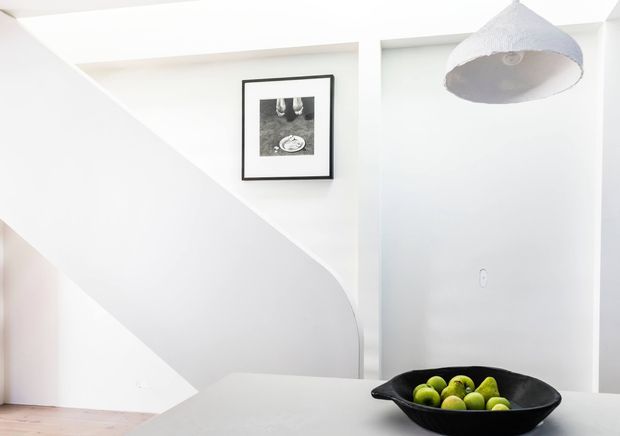 Bright minimalist interior featuring a white sculptural stair or curved partition wall, pale timber flooring, a simple framed monochrome artwork, a white pendant light, and a dark bowl of green fruit on a light table in the foreground.