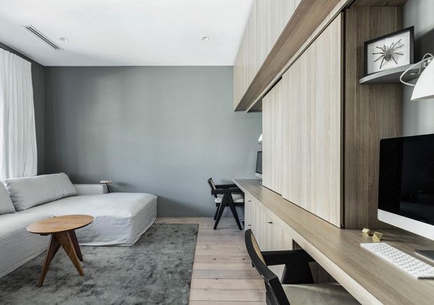 A compact, contemporary living room and integrated home workspace. The room features a light sectional sofa, round timber coffee table, grey rug, grey painted wall, pale timber flooring, and extensive dark timber built-in cabinetry with a desk, shelving, computer monitor and concealed storage. Sheer curtains and recessed ceiling lighting contribute to a clean, refined interior.