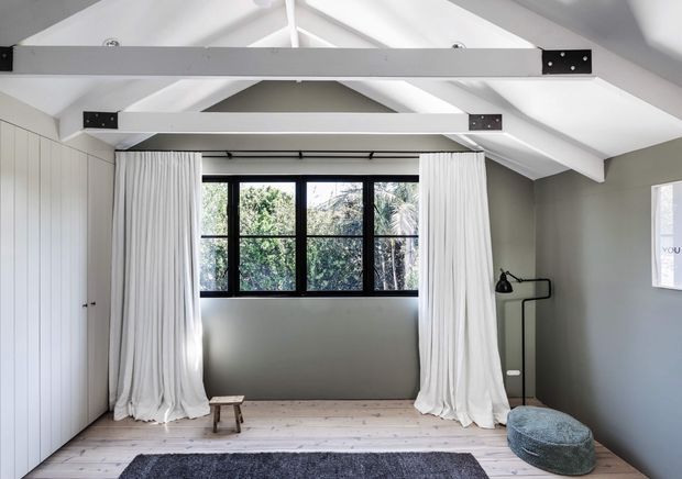 Bright attic-style bedroom with a pitched white ceiling and exposed painted structural beams, pale timber flooring, built-in white wardrobe cabinetry, black-framed multi-pane window, full-height white curtains, and a simple black floor lamp. The room has a calm, minimalist character with a small stool, textured rug, and low upholstered chair.