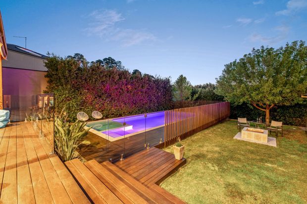 A landscaped residential backyard viewed from a timber deck, featuring a long illuminated swimming pool enclosed by frameless glass fencing, timber screening, lush hedging, and a small outdoor seating area beneath a mature tree. The scene has warm evening lighting with purple pool illumination and a contemporary outdoor entertaining character.