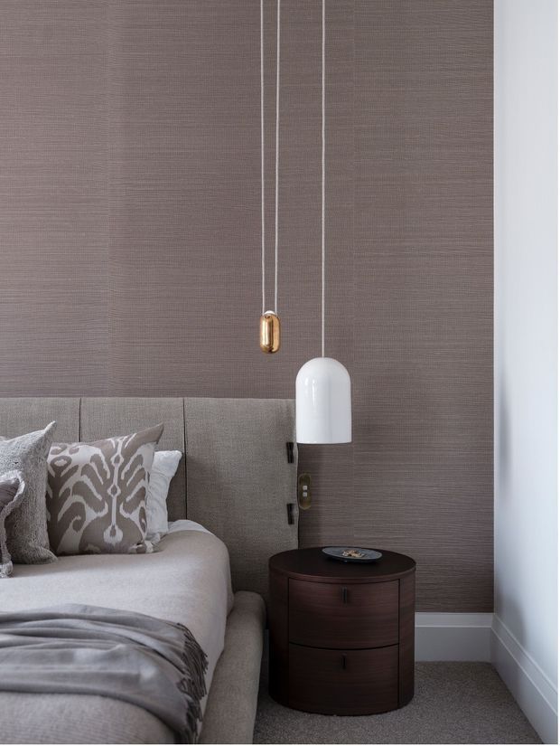 A refined bedroom corner featuring a fully upholstered, vertically panelled headboard, taupe textured wall covering, soft neutral bedding, patterned cushions, and a dark timber round bedside table. A white pendant light and small brass pendant hang beside the bed, creating a sophisticated layered lighting arrangement. The palette is muted and warm with contemporary detailing.