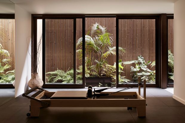 A refined indoor exercise room featuring a wooden Pilates reformer positioned in front of full-height dark-framed glazing. Vertical timber screening and lush planting are visible beyond the windows, creating a calm connection to an enclosed garden. The room has warm neutral walls, a dark tiled floor, sculptural woven vessels, and soft natural light.
