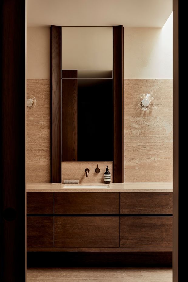 A refined bathroom vanity viewed straight on, featuring a dark timber floating cabinet with integrated drawers, a pale stone benchtop and basin, and a large vertically framed mirror. Warm beige stone or tile wall cladding covers the lower walls, with sculptural wall sconces on either side. The composition is symmetrical and softly lit, with a warm, luxurious contemporary feel.