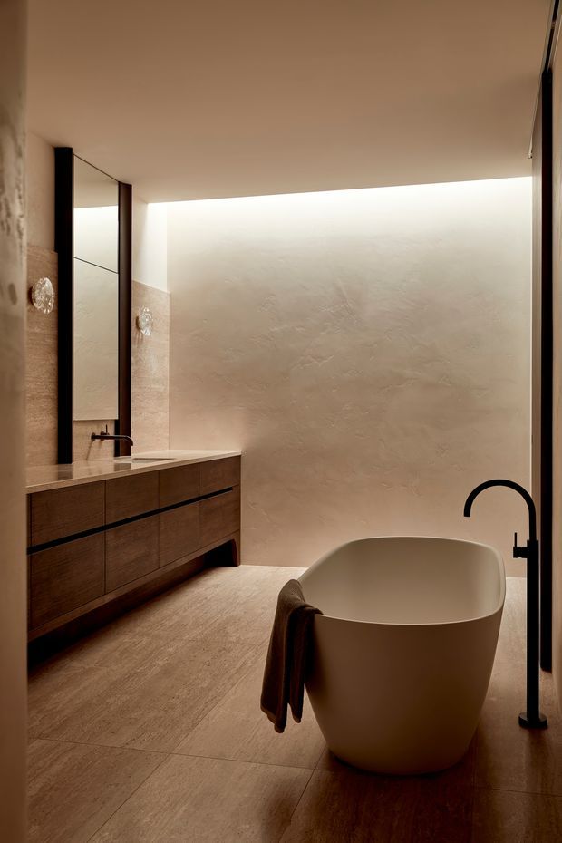 A warm, minimalist bathroom featuring a freestanding oval bathtub, a dark wall-mounted vanity with integrated basin, large vertical mirror panels, black tapware, and softly textured beige plaster walls. Large-format beige stone-look floor tiles and concealed linear lighting create a calm, refined atmosphere.